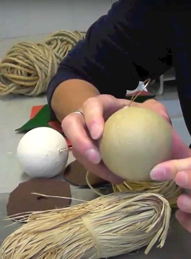 supplies for showing how to make a scarecrow craft | holding paper mache ornament with a painted ornament in the background along with rope and there is raffia in the foreground 