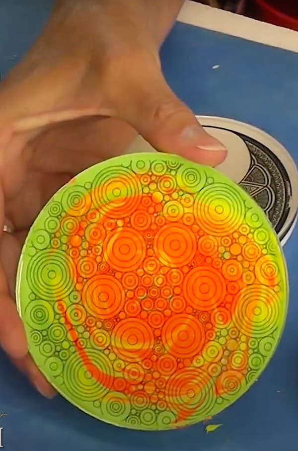 Holding up swirl mandala art coaster with orange and green neon colors reveal